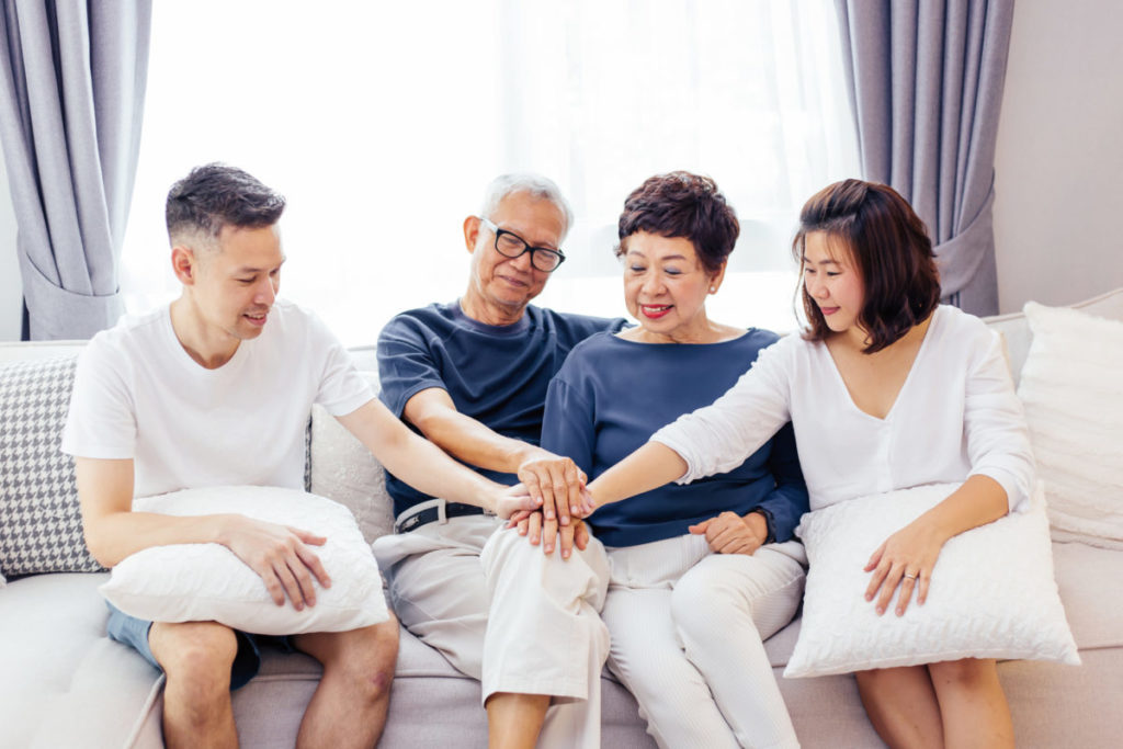 Asian family with adult children and senior parents putting hands together and sitting on sofa at home together. Family unity and cooperation concept