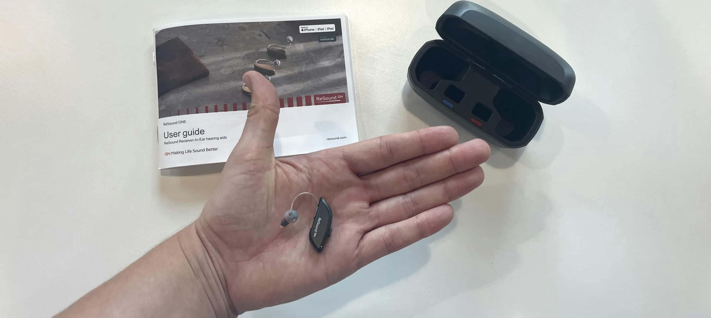 Resound hearing aid in outstretched hand with charging case and instruction manual