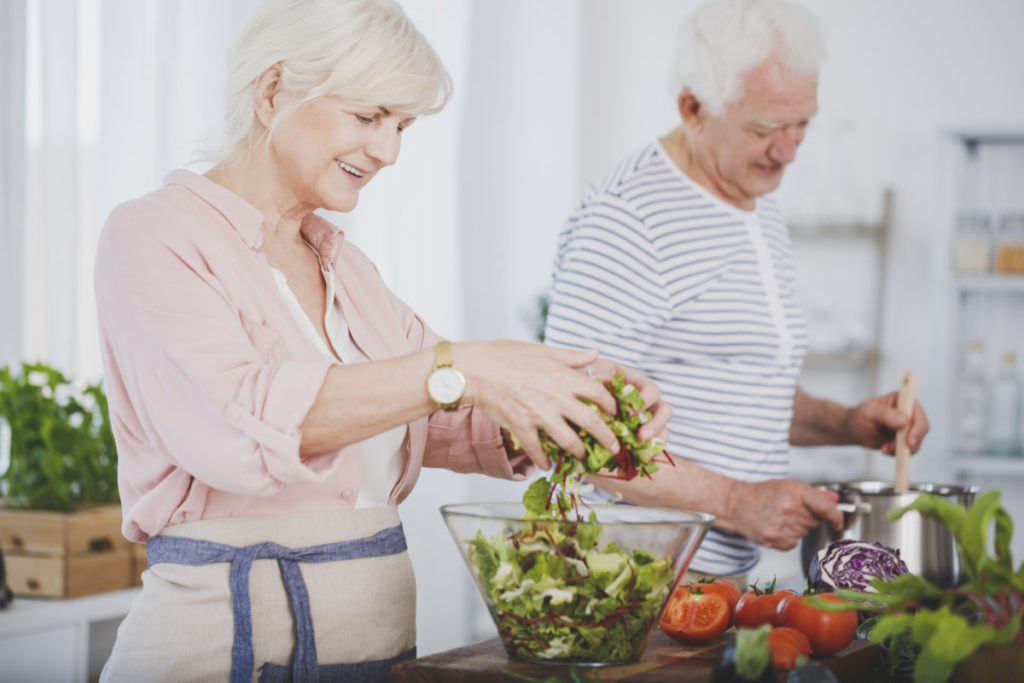 Older woman tossing a salad and her husband stirring food in a pot, cooking healthy together