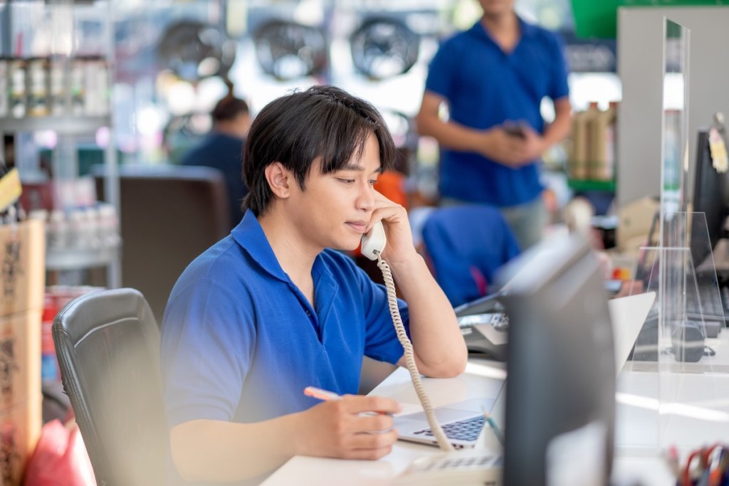 Asian man receive phone call from customer and stay in counter service area of automotive or garage shop with other worker work in the background