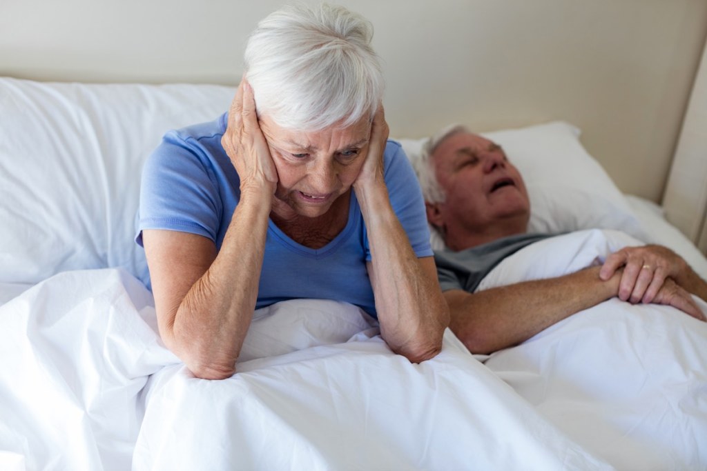 Senior woman getting disturbed with man snoring on bed in bedroom